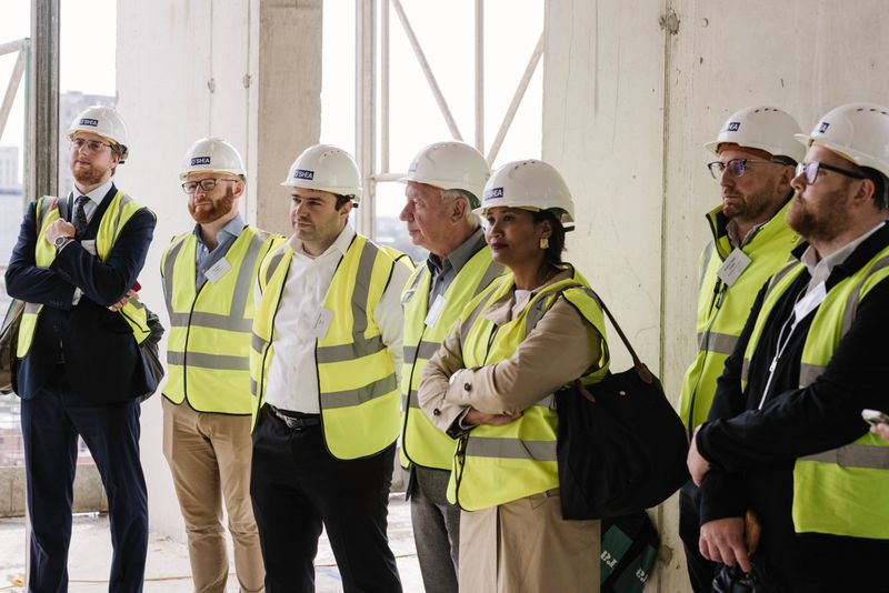 Group of people wearing white hard hats and high-visibility safety vests standing inside a partially constructed building.