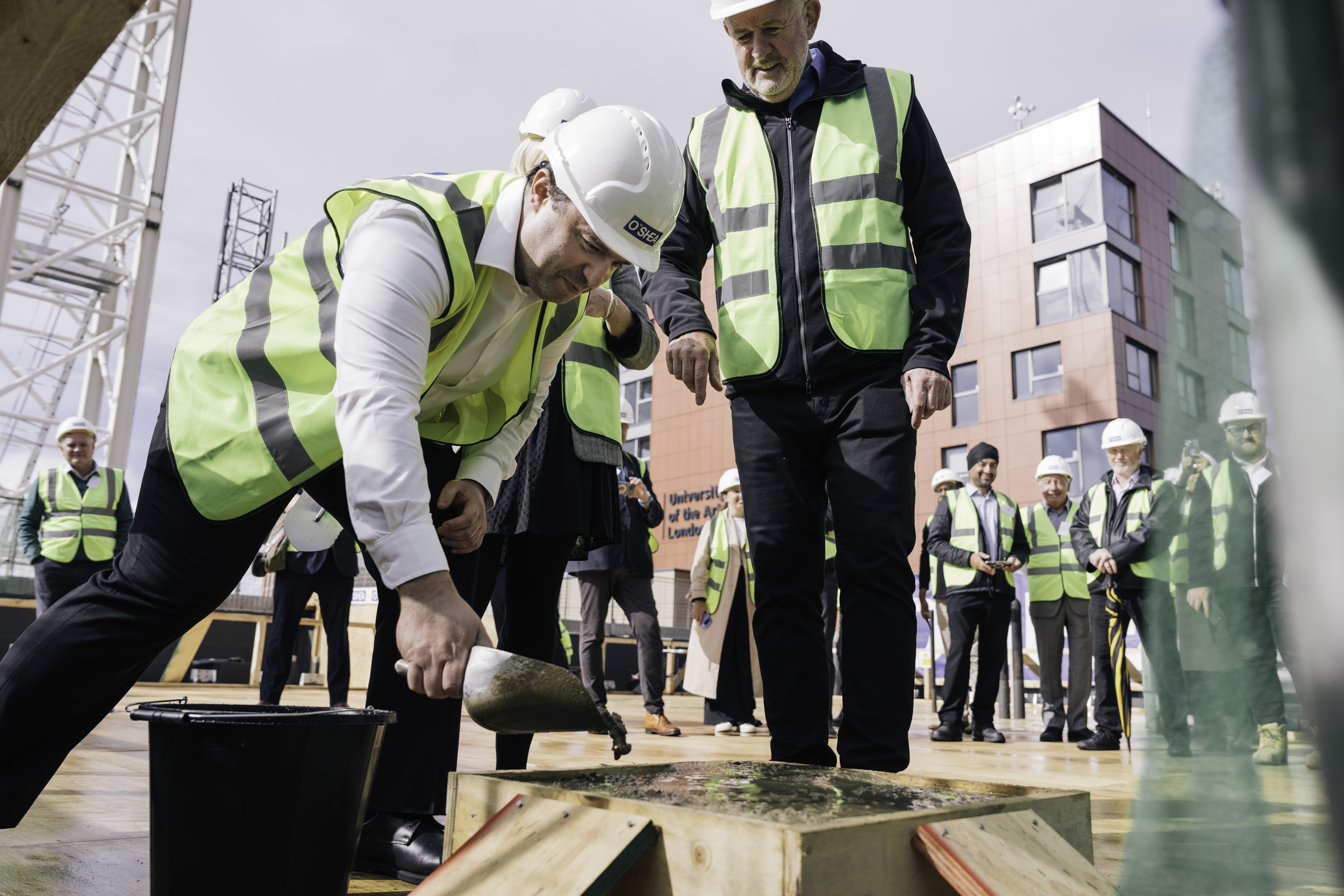 Person in a high-visibility vest and hard hat pouring concrete during a construction ceremony.